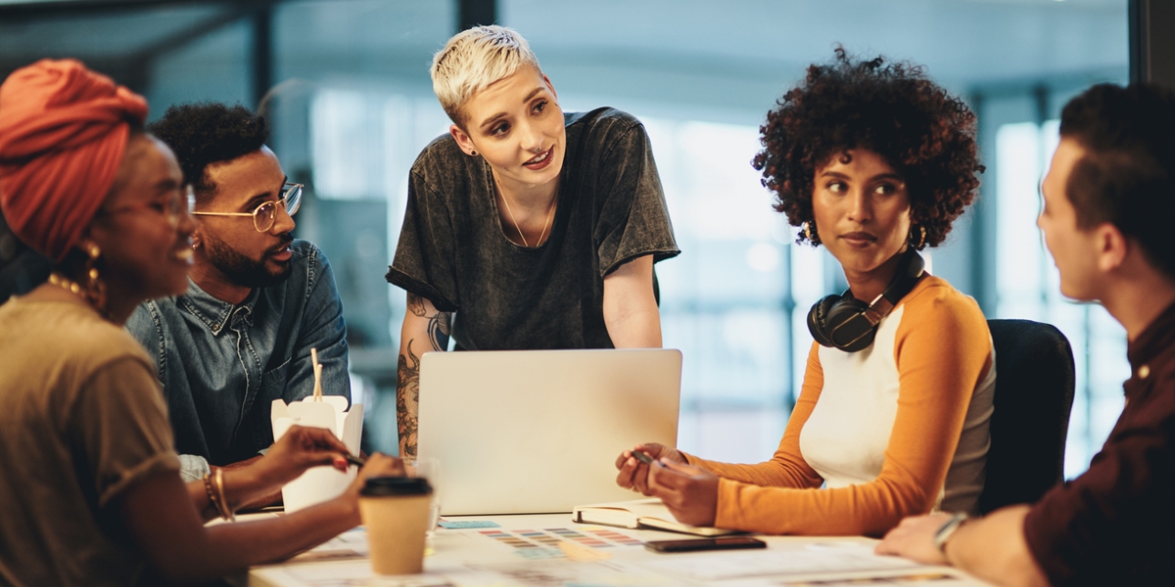 A group of five young professionals around a table: a Black woman with red turban, a black man wearing a denim shirt, a white woman with bleached hair and tattooed arms stands, a Black woman with headphones around her neck, and a white man who can be