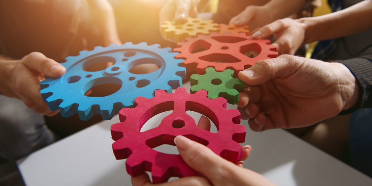 Six hands holding four cogs of various sizes together