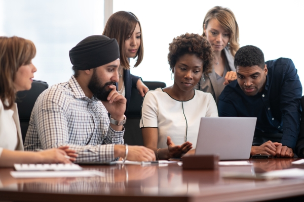 Five colleagues gather around a laptop. They consist of a white woman with brown hair wearing a cream jacket, an Asian man wearing a navy turban and check shirt, a Black woman with large hooped earrings and cream jacket, and a Black man with short ha