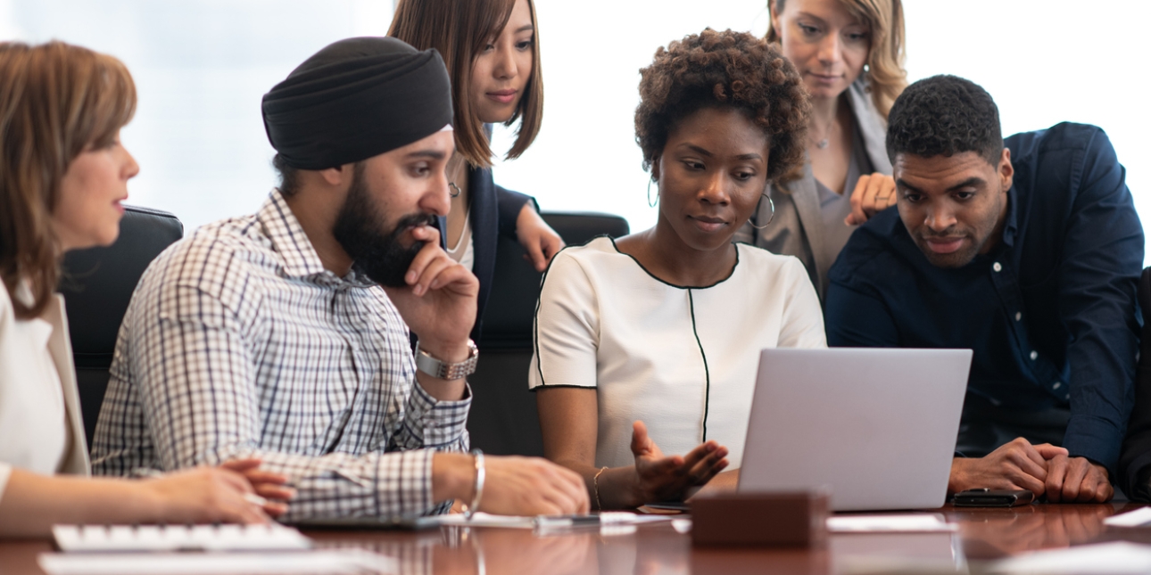 Five colleagues gather around a laptop. They consist of a white woman with brown hair wearing a cream jacket, an Asian man wearing a navy turban and check shirt, a Black woman with large hooped earrings and cream jacket, and a Black man with short ha