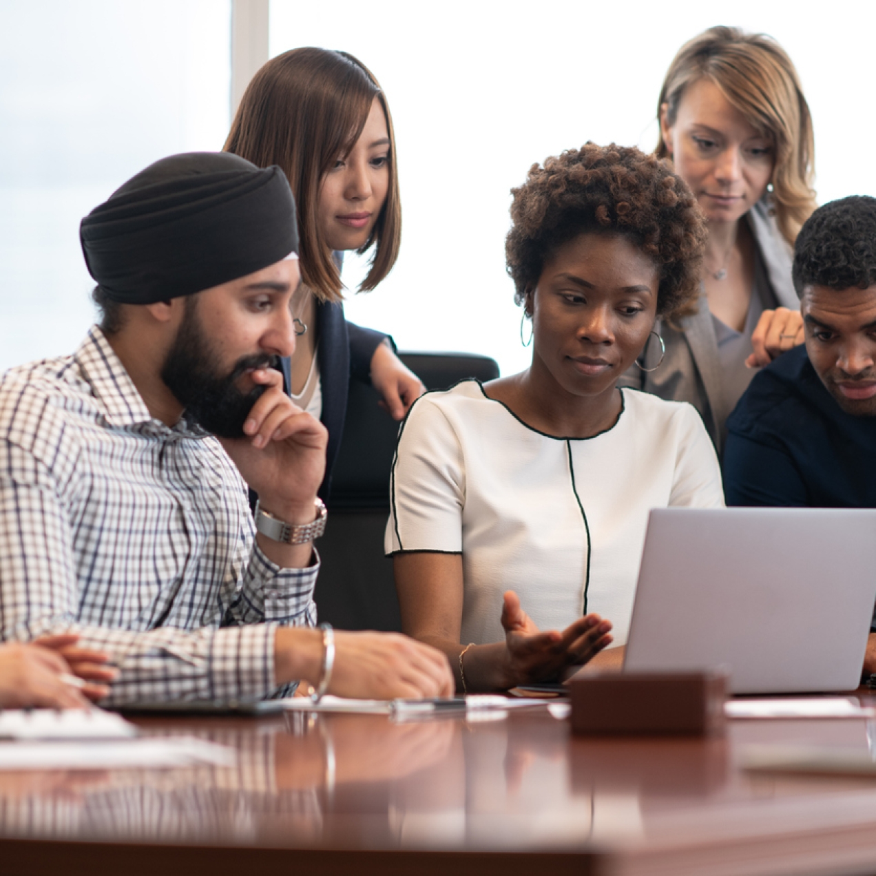 Five colleagues gather around a laptop. They consist of a white woman with brown hair wearing a cream jacket, an Asian man wearing a navy turban and check shirt, a Black woman with large hooped earrings and cream jacket, and a Black man with short ha