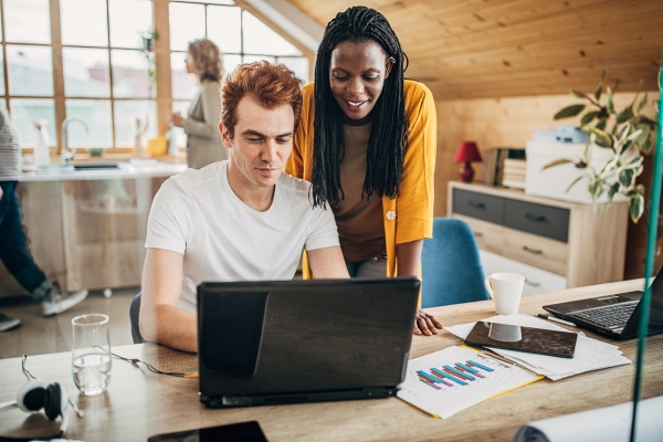 A Black woman with long braided hair and wearing an orange cardigan smiles while standing over the shoulder of a white male colleague. He has ginger hair and wears a white t-shirt, and is sat showing her his laptop screen. The background shows a brig