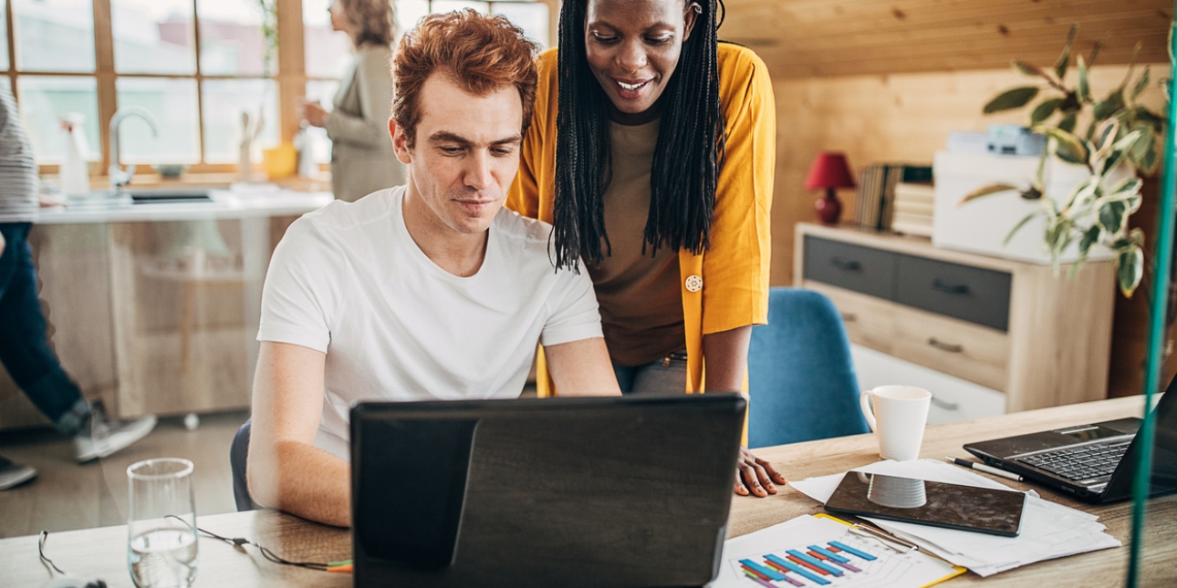 A Black woman with long braided hair and wearing an orange cardigan smiles while standing over the shoulder of a white male colleague. He has ginger hair and wears a white t-shirt, and is sat showing her his laptop screen. The background shows a brig