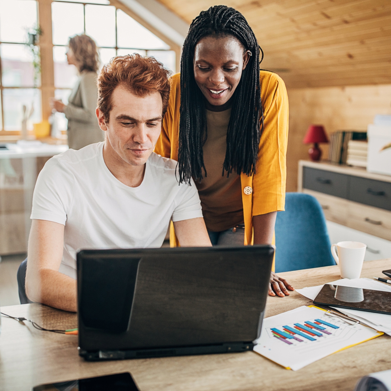 A Black woman with long braided hair and wearing an orange cardigan smiles while standing over the shoulder of a white male colleague. He has ginger hair and wears a white t-shirt, and is sat showing her his laptop screen. The background shows a brig