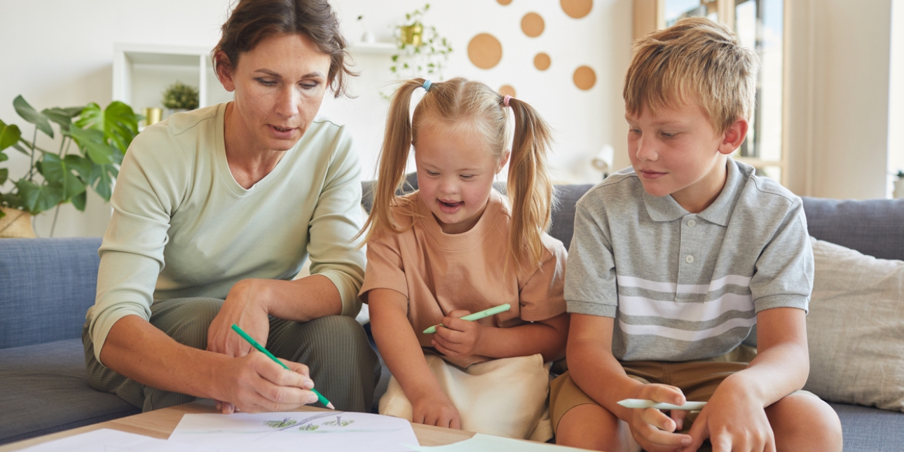 A mother, daughter and son are drawing together on a coffee table while sat on a sofa. The mother is white with dark hair and green top. The daughter is white, has blonde hair and has Down's syndrome. She wears a peach dress and is smiling. The son i