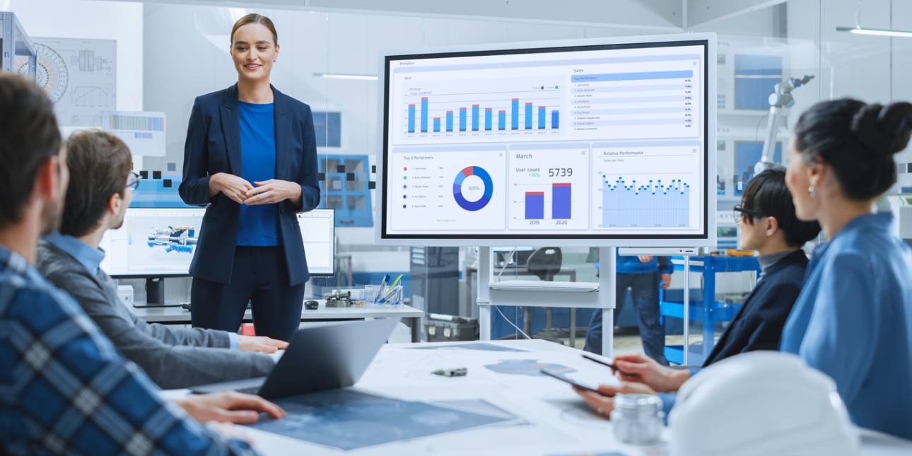 A woman, who is white and wears a dark suit and blue top, presents sales data on a screen to two men and two women sat around a meeting table. Behind her is a glass wall which shows a modern factory