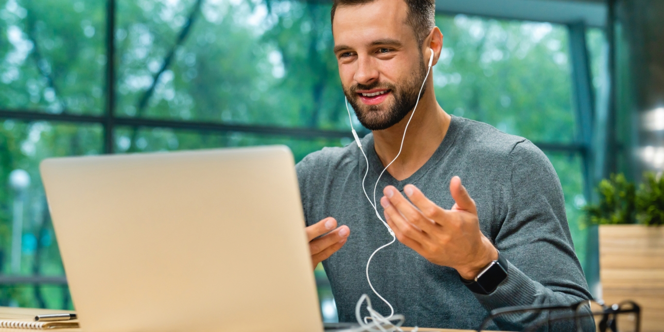 A white man with dark hair and beard, wears a grey sweatshirt while sat at a desk. He is mid conversation speaking on his laptop screen and wears white earphones