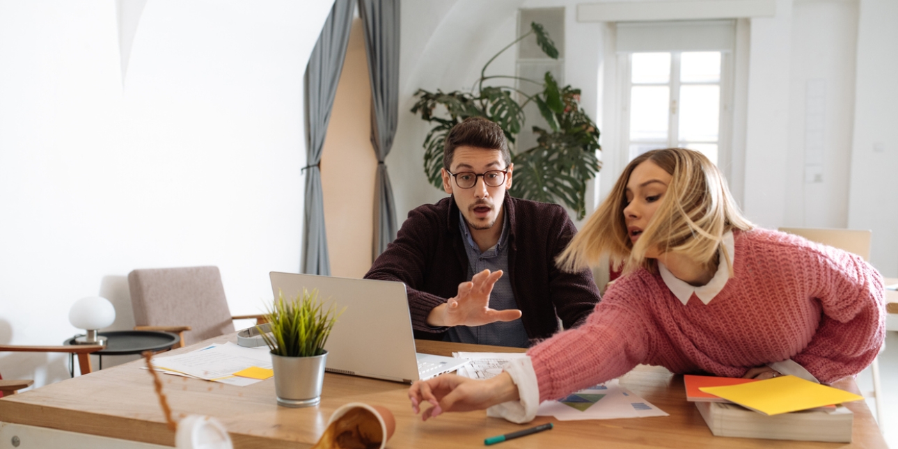 A white woman reaches across the desk in a vain attempt to rescue a cup of coffee which has been knocked over, spilling liquid everywhere. On the left is a white man with a horrified expression reaching out to help