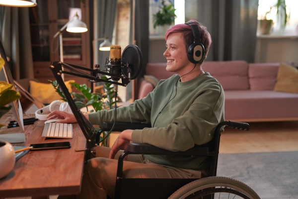 A white woman with short pink hair and wearing large headphones is sat in a wheelchair in front of a desk with podcasting equipment including a microphone