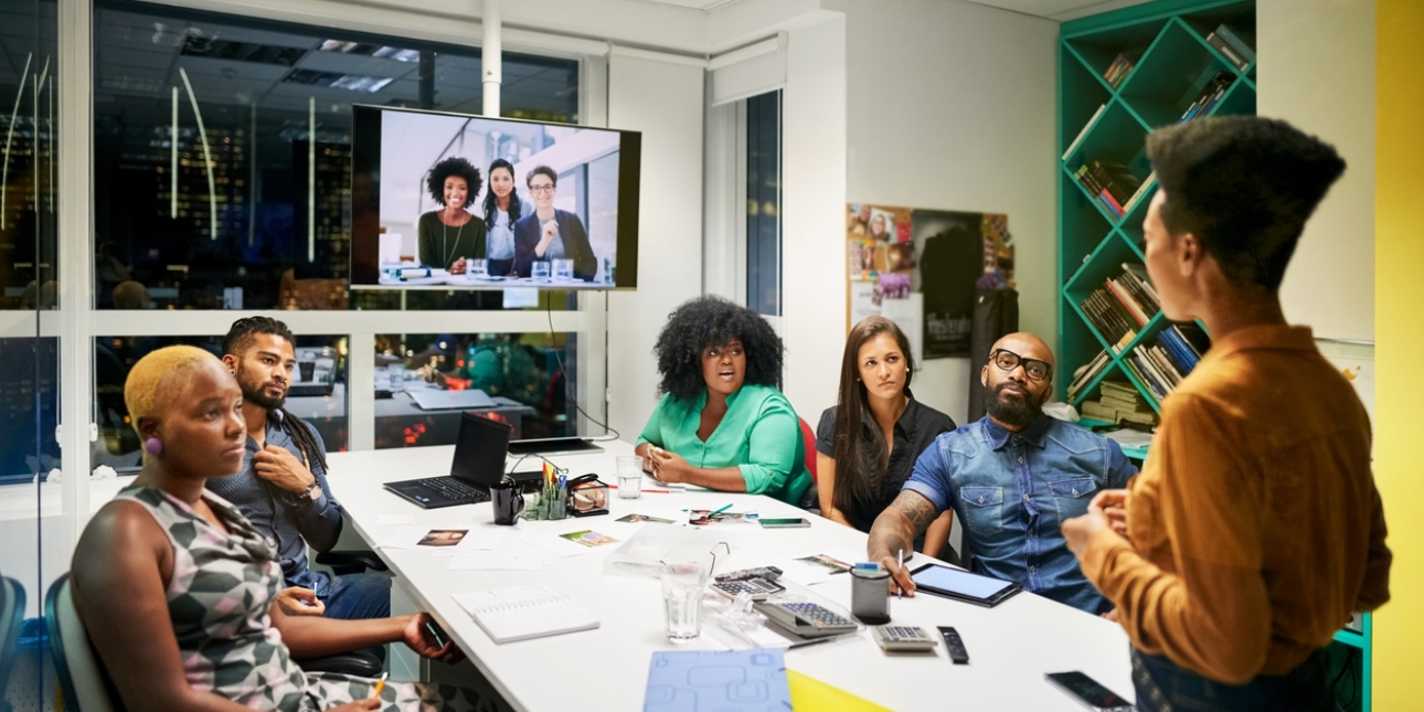 A group of colleagues sat around a board table look to another who is stood up talking. Three colleagues have joined on a video screen.