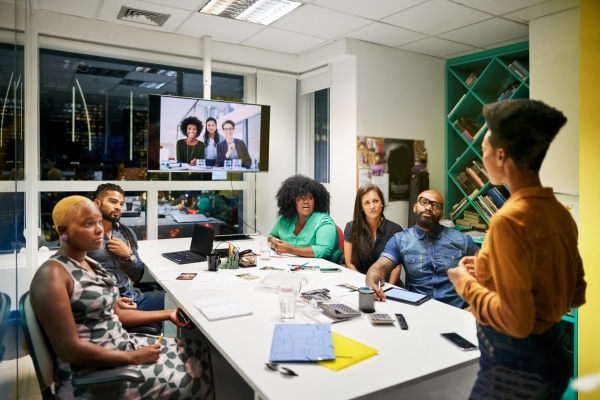 A group of colleagues sat around a board table look to another who is stood up talking. Three colleagues have joined on a video screen.
