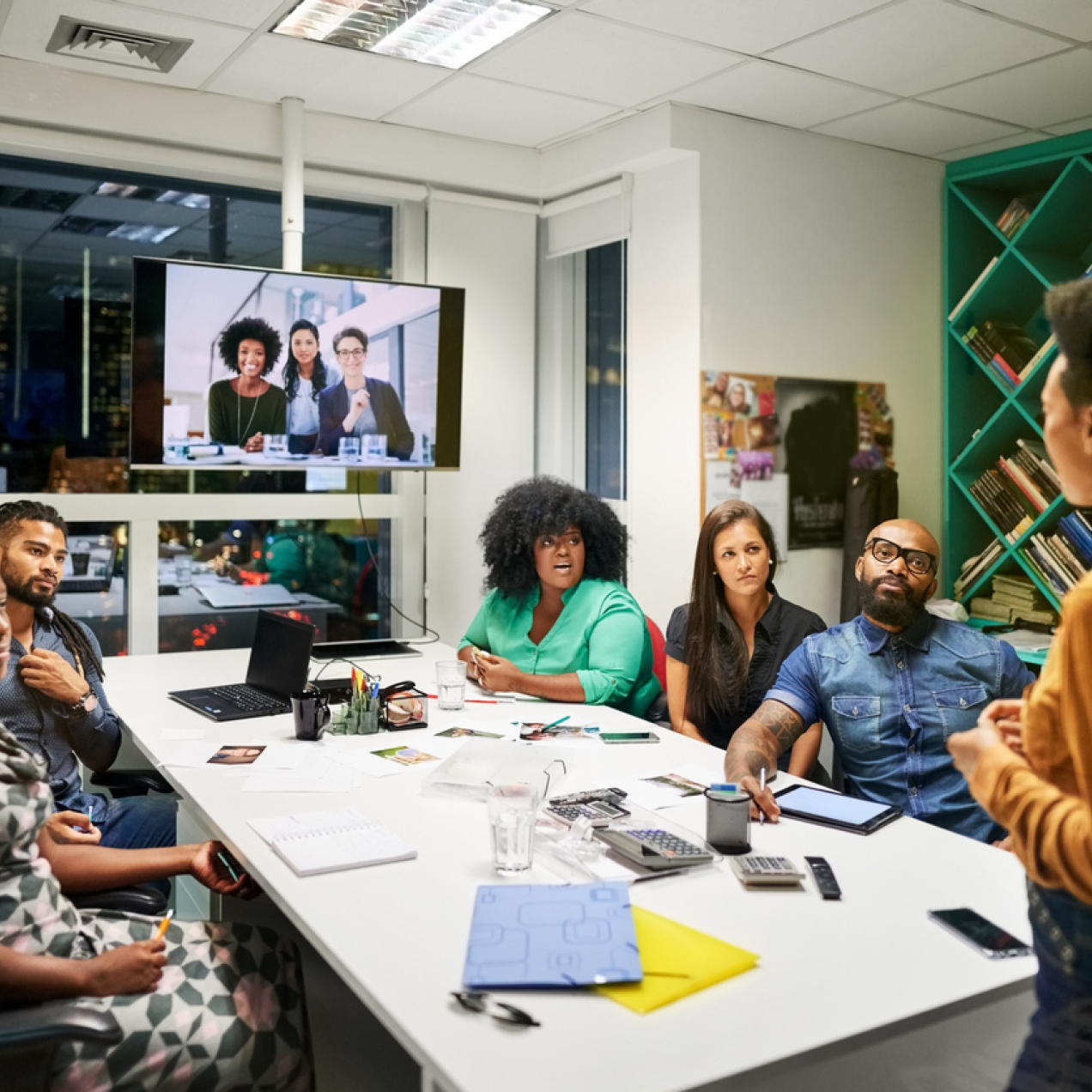 A group of colleagues sat around a board table look to another who is stood up talking. Three colleagues have joined on a video screen.