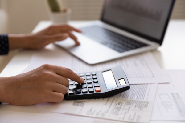 Close up of fingers on a calculator on top of bills. The other hand is typing on a laptop.
