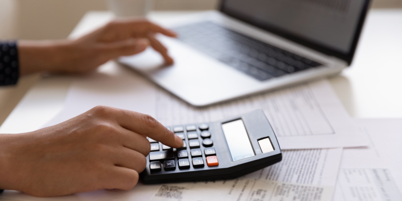 Close up of fingers on a calculator on top of bills. The other hand is typing on a laptop.