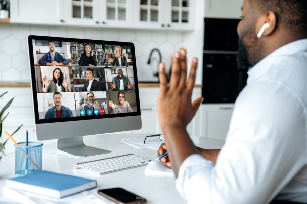 A Black man in a grey shirt sits in his kitchen waving to nine colleagues on his computer screen. The colleagues are male and female and of different ethnicities. They are in different locations and smiling or waving