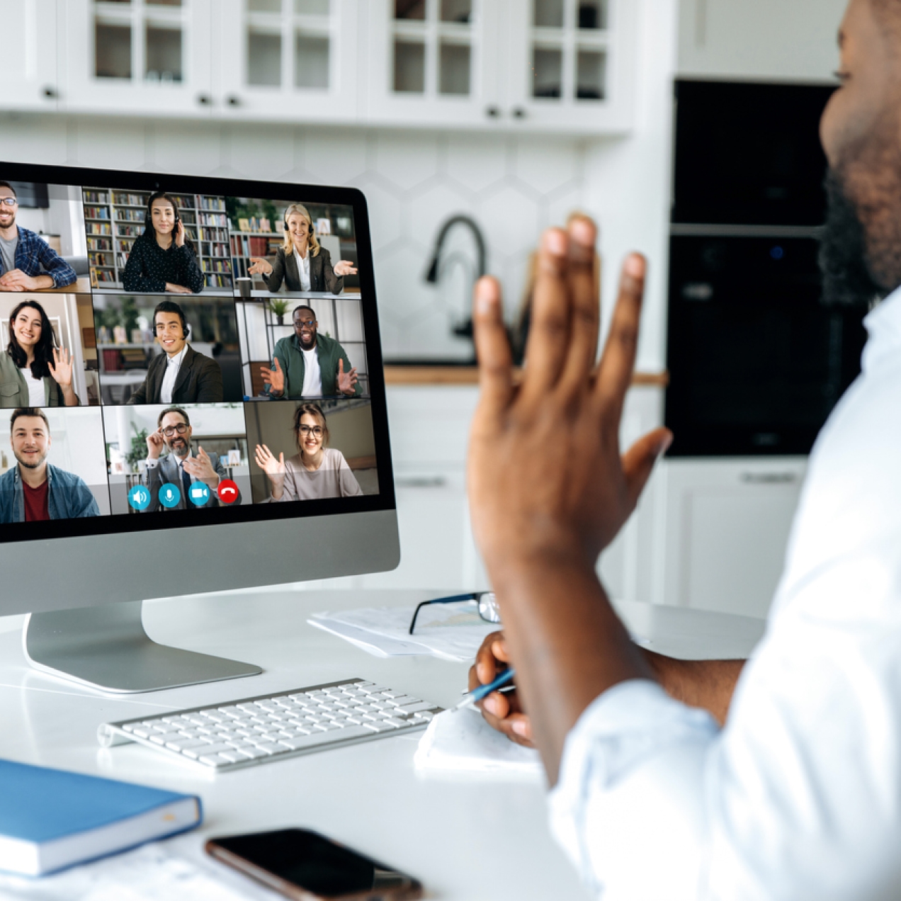 A Black man in a grey shirt sits in his kitchen waving to nine colleagues on his computer screen. The colleagues are male and female and of different ethnicities. They are in different locations and smiling or waving