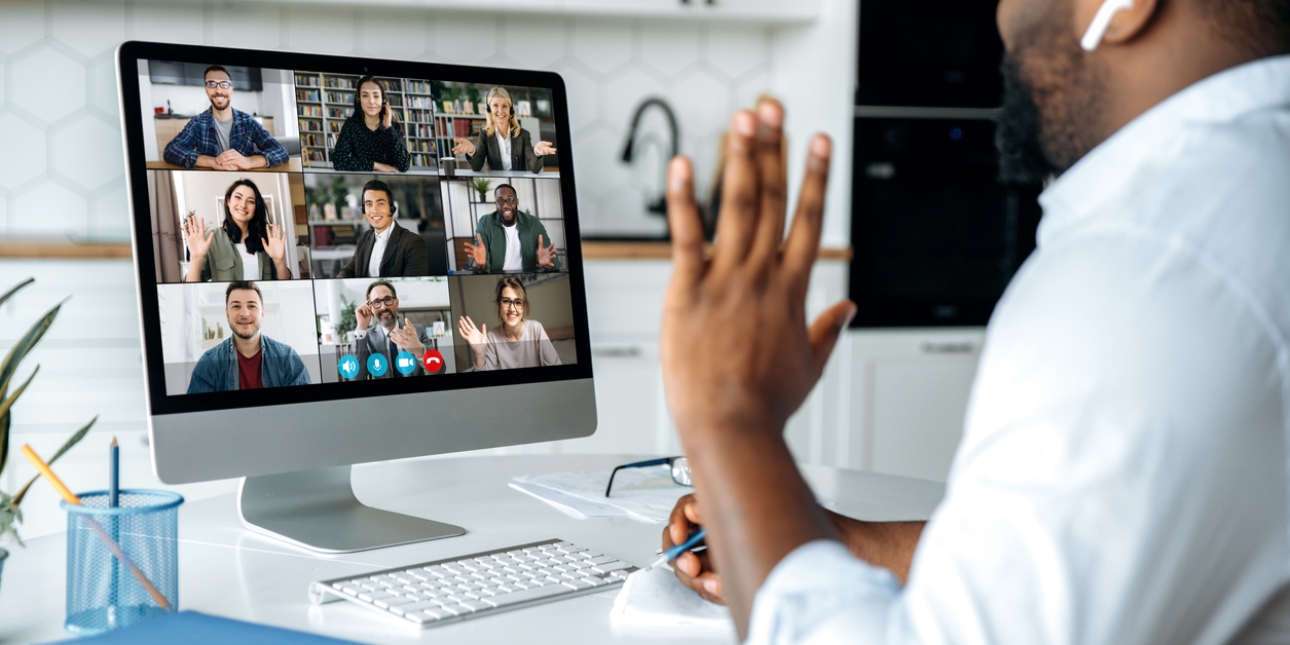 A Black man in a grey shirt sits in his kitchen waving to nine colleagues on his computer screen. The colleagues are male and female and of different ethnicities. They are in different locations and smiling or waving