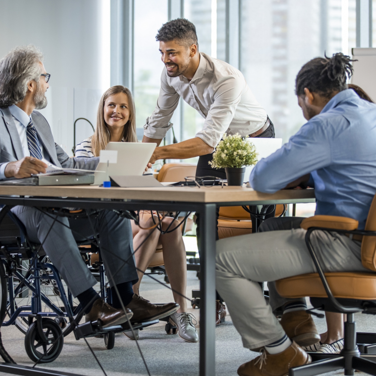 Five business colleagues sat around a table. On the left is a man sat in a wheelchair, next to him a woman smiling and stood over the desk is man also smiling. Another man and woman have their backs to camera.