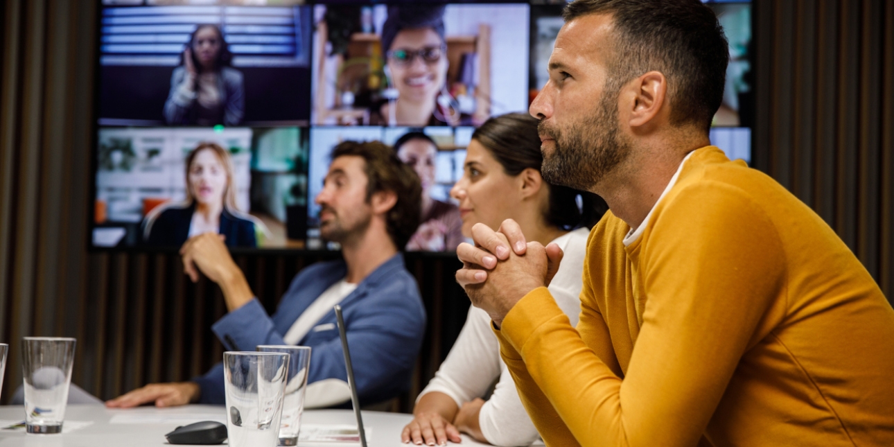 Colleagues sat around a table. Behind them is a screen with nine others joining on a video conference.