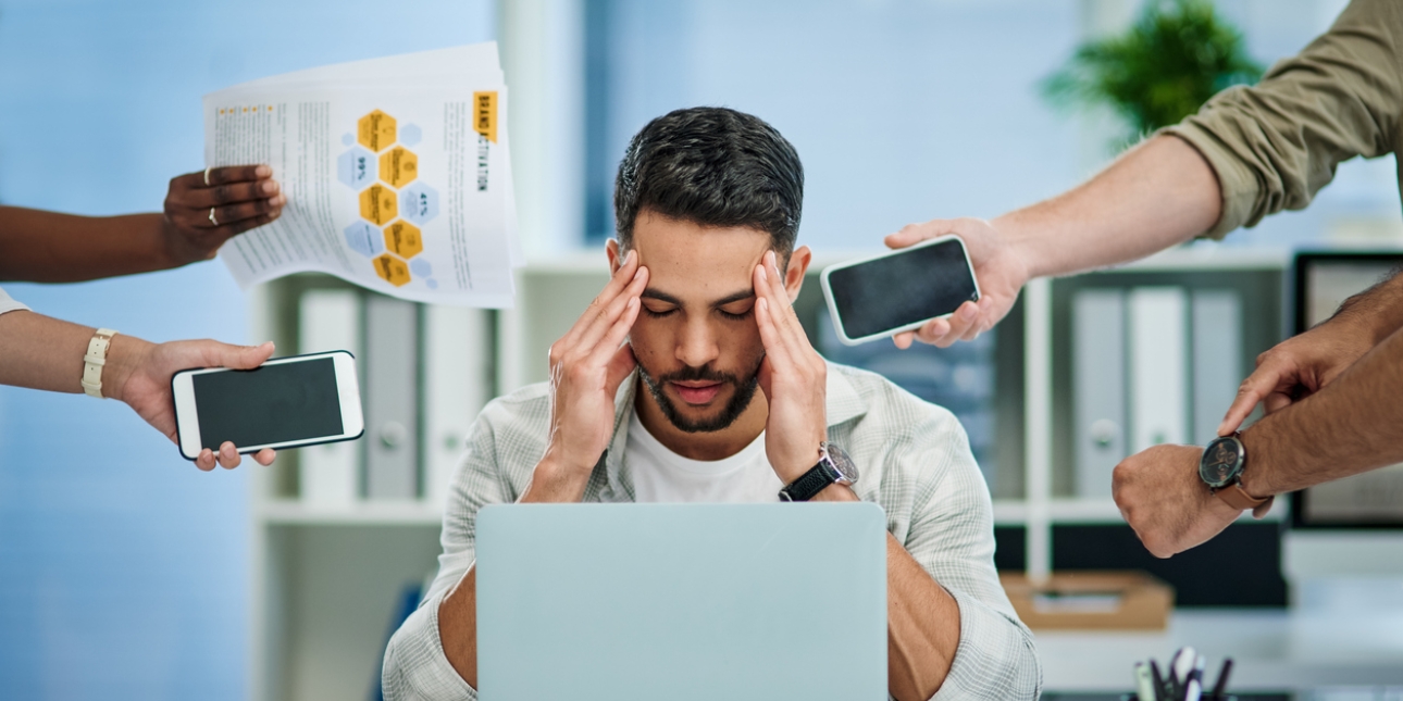 A white man with short dark hair and beard holds his head in his hands in front of his laptop. Surrounding him are unidentified hands holding mobile phones, paperwork and pointing at a wristwatch