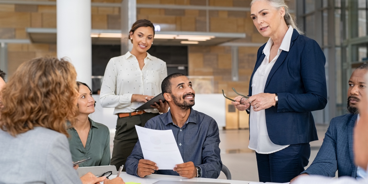 Colleagues sat smiling around a table on which sits a laptop and paper charts. An Asian man in a blue shirt looks up at a white female colleague with grey hair who is standing and holding spectacles. Behind him is an Asian female colleague. Around th