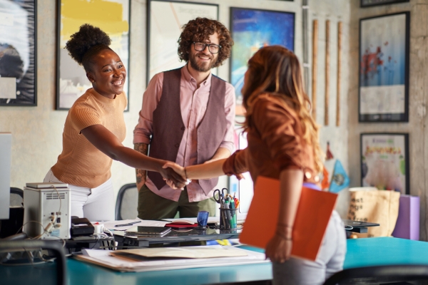 A smiling Black woman wearing a shortsleeved beige top, extends her hand over the desk to another woman, who is white and wearing a brown top. A white man with glasses and a beard, is also stood behind the desk smiling. He wears a pink shirt and mauv