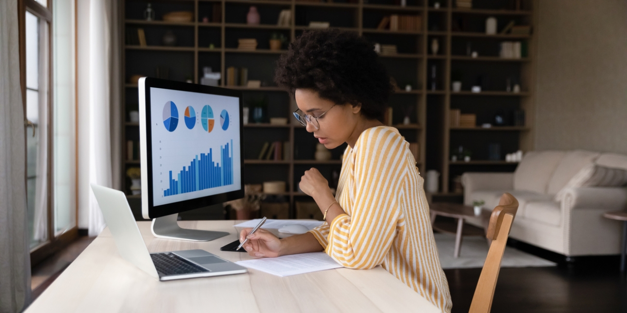 A woman at her desk, writing notes, with pie charts and graphs visible on her computer screen
