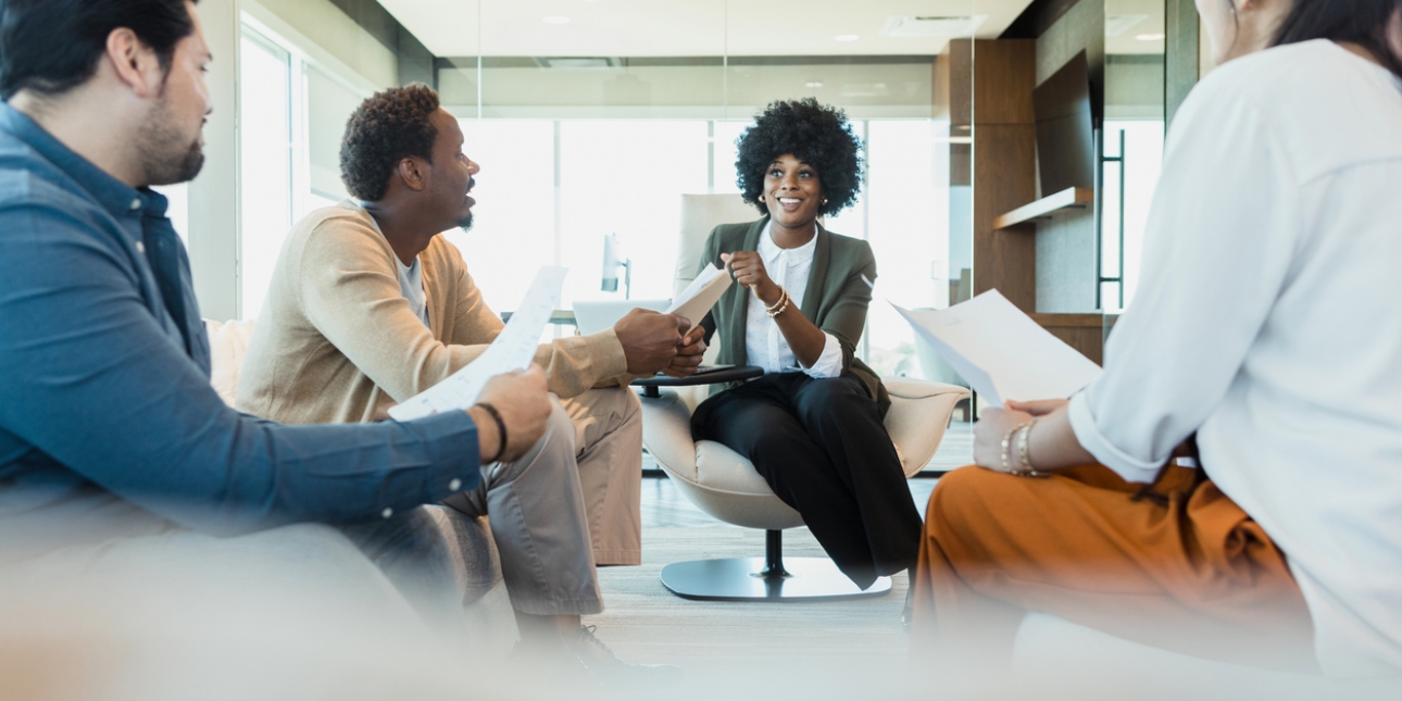 A Black woman CEO wearing a dark green jacket and white shirt, sits in a large chair addressing colleagues: a Black man in cream cardigan, an Asian man in blue shirt and a woman with long dark hair whose face can't be seen but wears a orange trousers