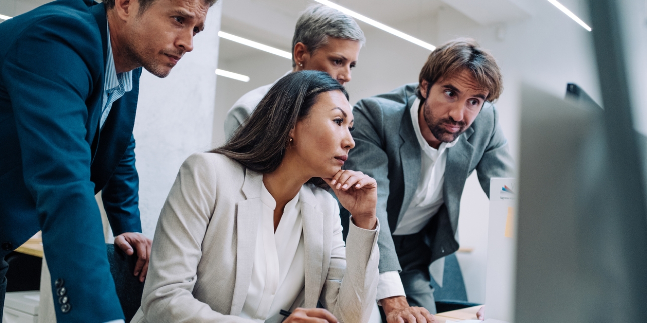 Three men stand over a colleague at her computer. All look serious. All are wearing business jackets.