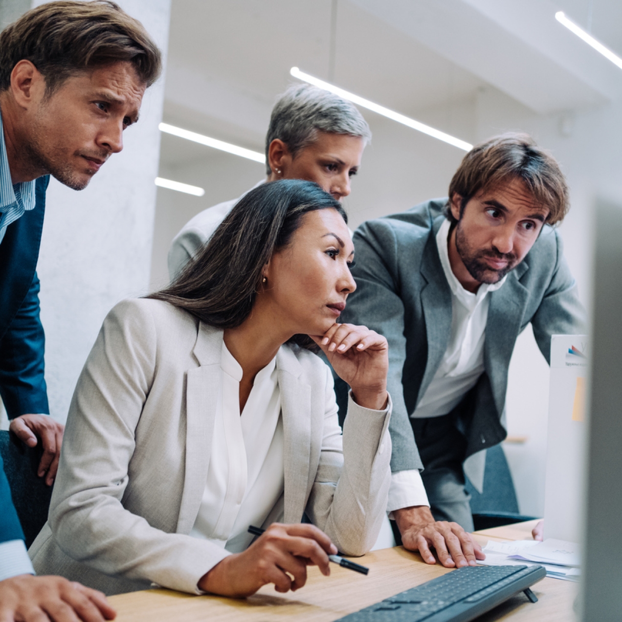Three men stand over a colleague at her computer. All look serious. All are wearing business jackets.