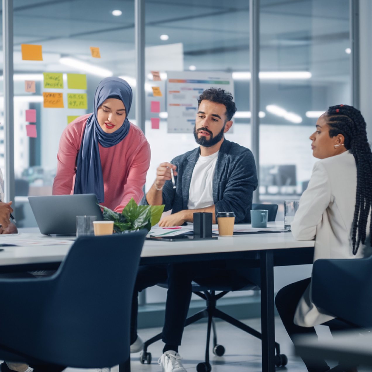 A woman in a blue hijab and pink jumper is stood pointing to a laptop while two male and one female colleague are sat around a board table