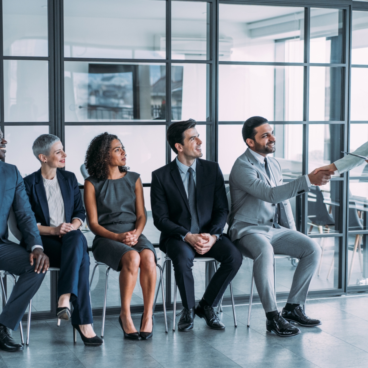 Five job candidates sit in a line against a glass office wall. There are two women and three men of different ethnicities. The man on the right shakes hand with the woman recruiter who is standing. All are smiling.