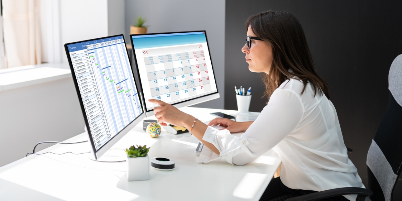 Side profile of a white woman with shoulder length dark hair, wearing a white shirt. She is sat at a desk in front of two monitors. The left hand monitor displays a Gantt chart, the right hand monitor shows a calendar. The walls are white and black.