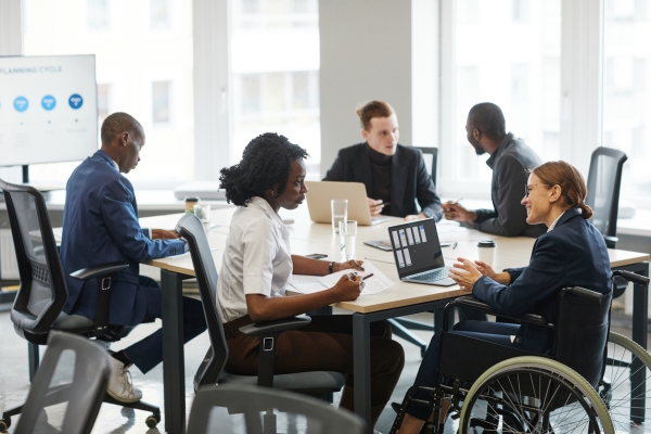 Five business people around a board table in a bright room. In the foreground a black woman in a white shirt and brown trousers is sat speaking to a woman colleague in a navy blazer, who uses a wheelchair. She has a laptop open in front of her. In th