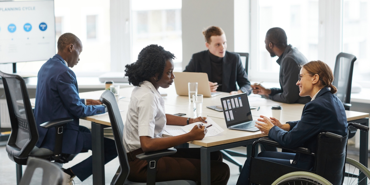 Five business people around a board table in a bright room. In the foreground a black woman in a white shirt and brown trousers is sat speaking to a woman colleague in a navy blazer, who uses a wheelchair. She has a laptop open in front of her. In th