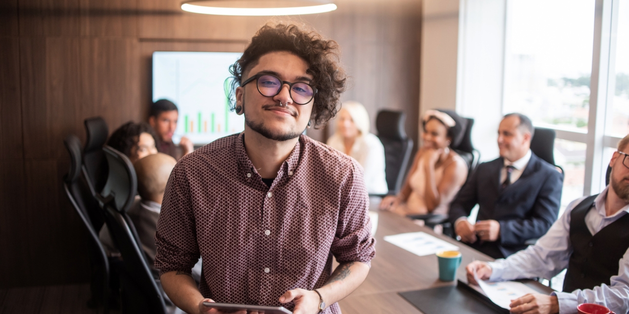 A trans white man with dark curly hair and glasses leans on a board table facing the camera. In the background are colleagues sat at the table.