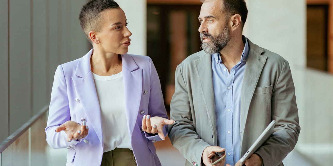 A woman and man in casual business dress in discussion while walking along a corridor. The man holds a phone and tablet.