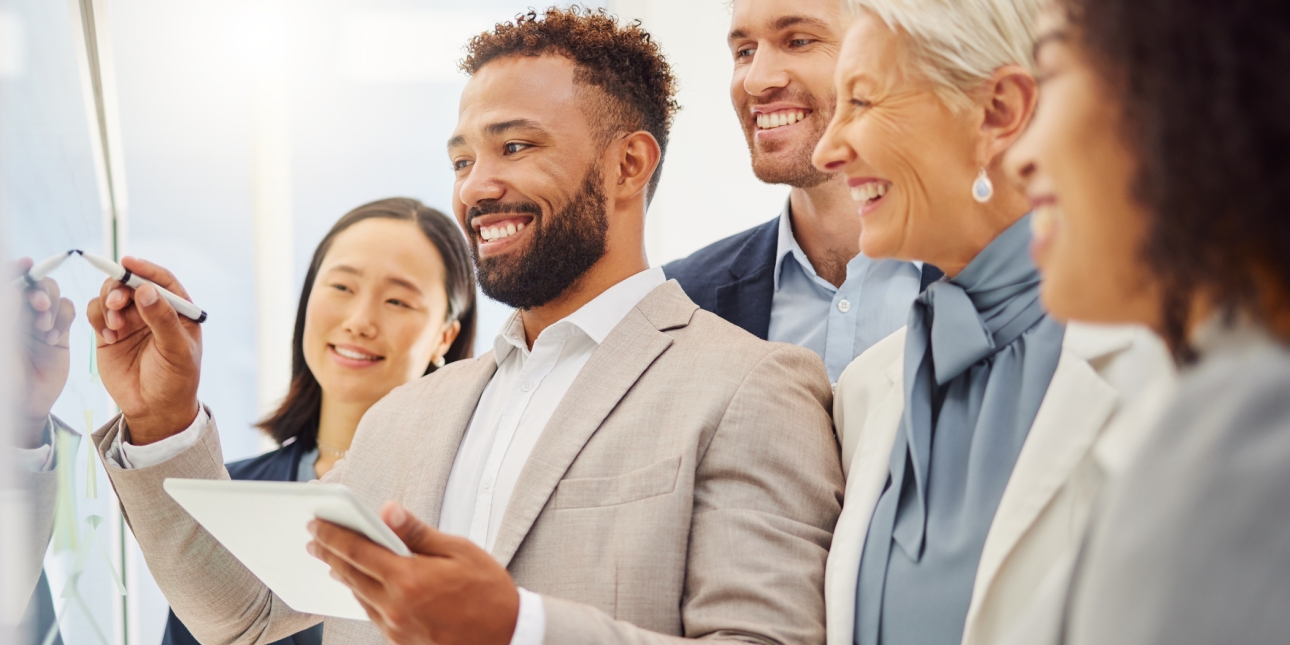 Six businesspeople gathered around a board. The man in the centre is holding a pen. All look happy.