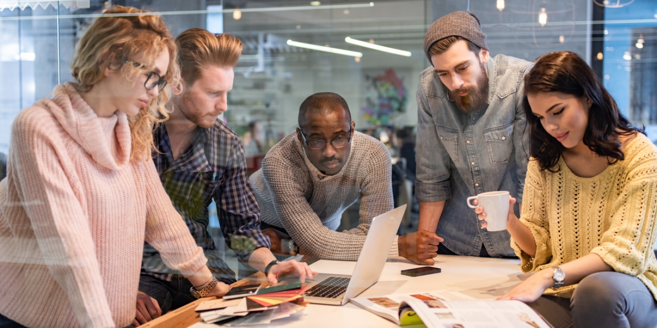 Five colleagues stood or sat around a desk in an office looking at a laptop, paperwork, a tablet, smartphones and a magazine. All are casually dressed. There are two white women, one Black man and two white men.