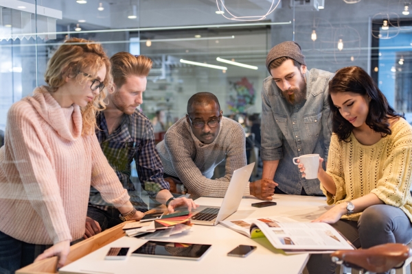 Five colleagues stood or sat around a desk in an office looking at a laptop, paperwork, a tablet, smartphones and a magazine. All are casually dressed. There are two white women, one Black man and two white men.