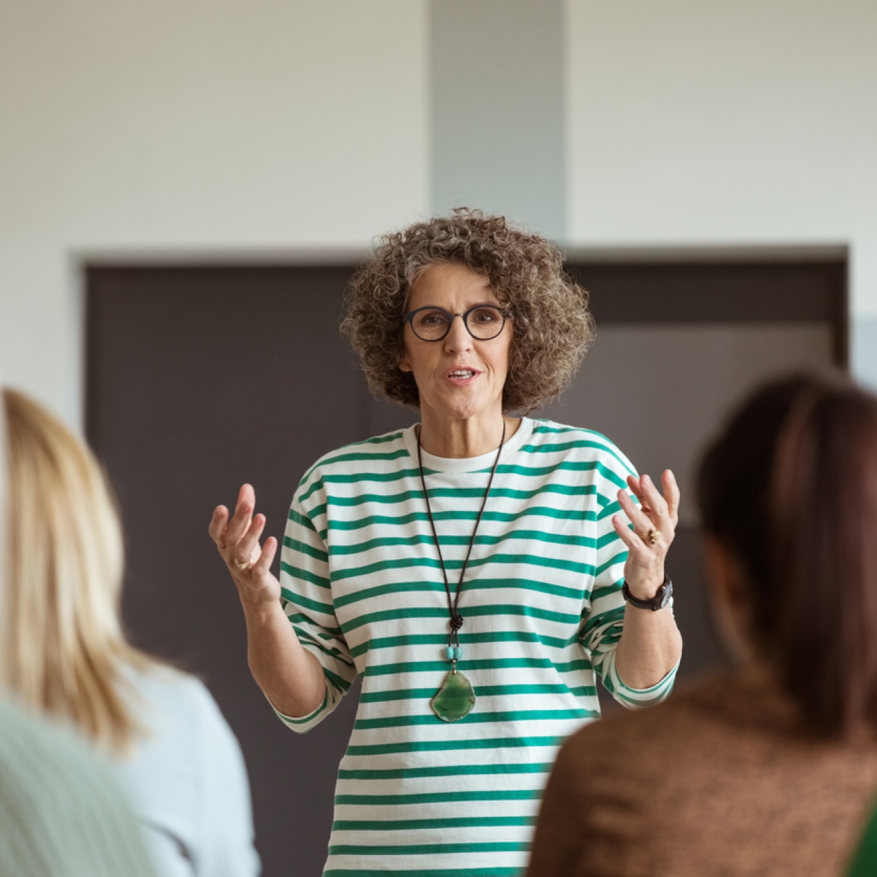 A woman, who is white with brown curly hair and glasses, stands in front of an audience. Her hands are raised to chest height. She wears a green and white striped top with a green stone pendant necklace.