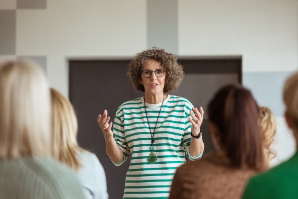 A woman, who is white with brown curly hair and glasses, stands in front of an audience. Her hands are raised to chest height. She wears a green and white striped top with a green stone pendant necklace.