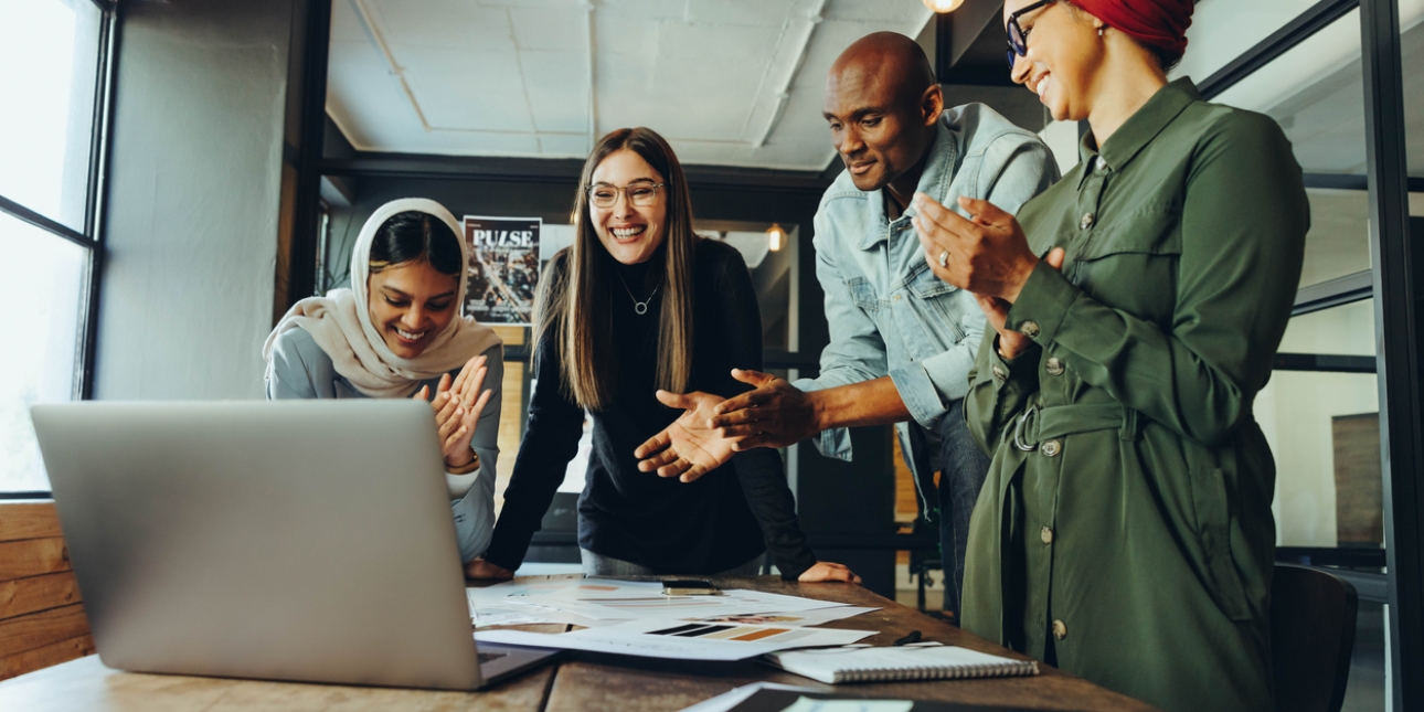 Four smiling colleagues stand around a laptop on a video call. They are an Asian woman wearing a hijab, a white woman wearing a necklace, a Black man wearing a denim jacket, and a Black woman wearing a red scarf in her hair