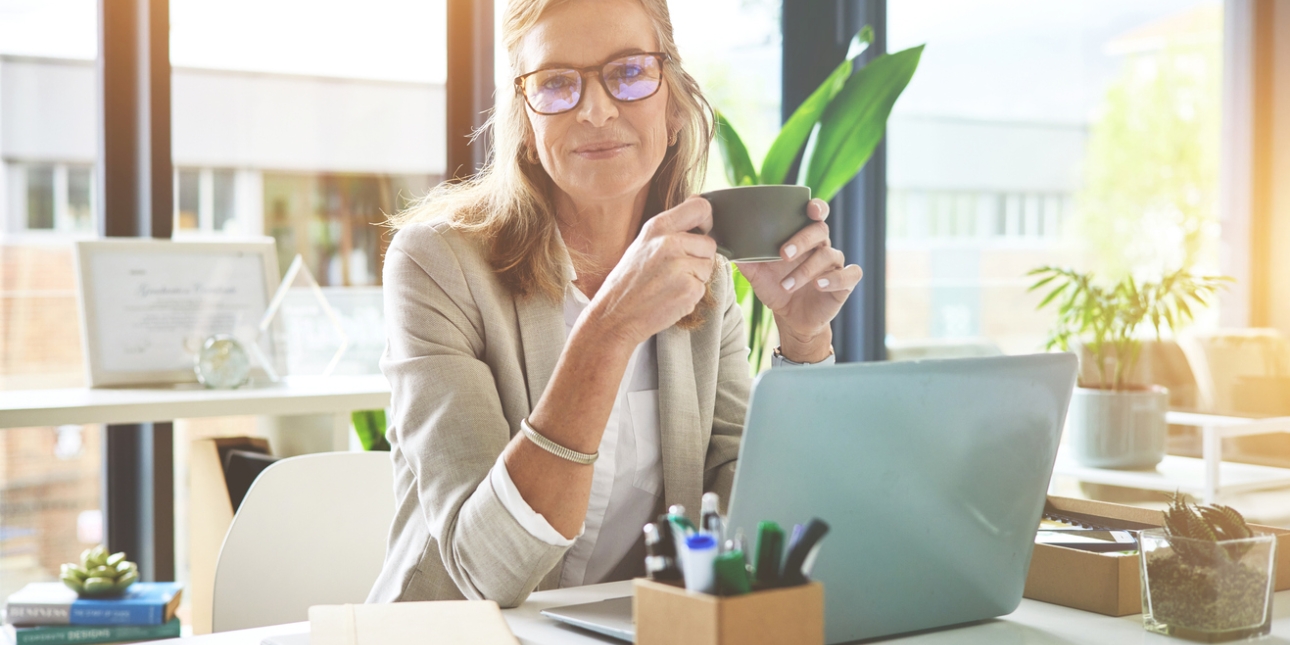 A woman with grey hair and glasses sits at a desk in front of a laptop while holding a cup of coffee