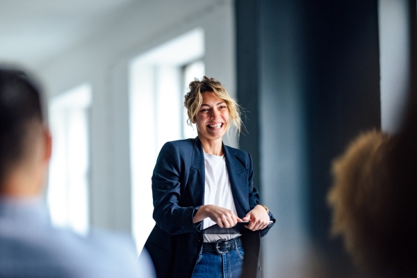 A white woman with blonde hair, smiles as she present to an audience. She wears a navy jacket, white top, blue jean, and is holding a tablet device.