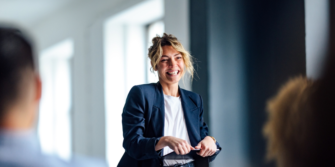 A white woman with blonde hair, smiles as she present to an audience. She wears a navy jacket, white top, blue jean, and is holding a tablet device.