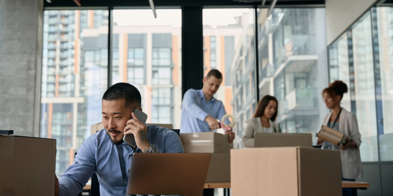 Office worker on the phone while surrounded by boxes. Three colleagues in the background packing boxes.