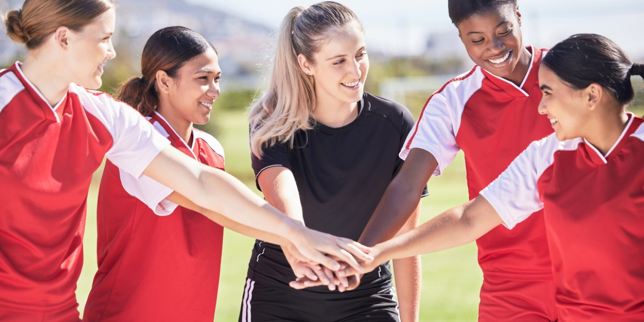 Four women football players in red jerseys and a woman referee in a black jersey place their hands on one another