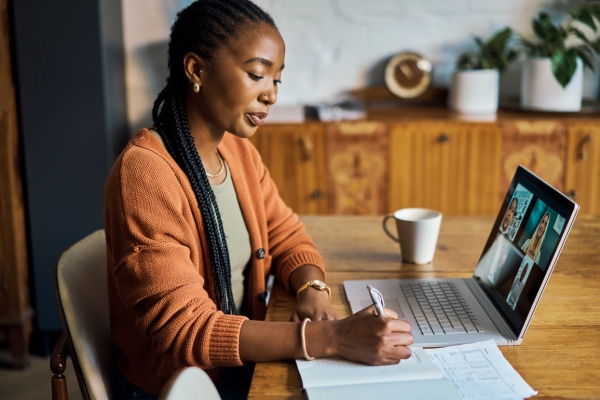 A Black woman with long hair and wearing an orange cardigan sits at a table writing notes in a book. In front of her is a laptop with four people on a webinar.