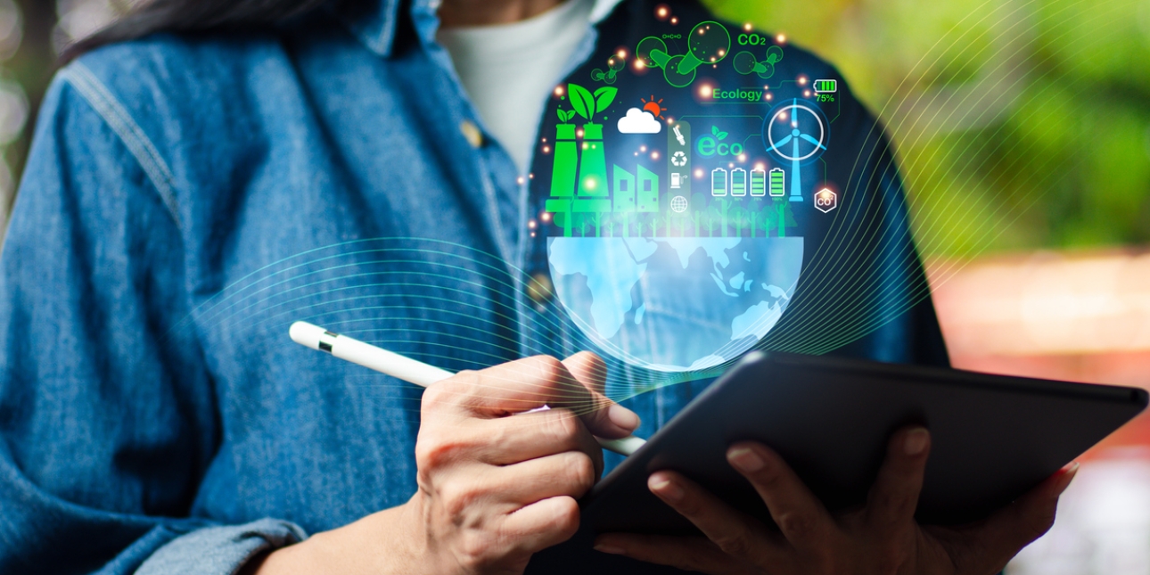 A woman in a denim shirt holds a tablet and stylus. A hologram image floats above the tablet featuring the southern hemisphere and various environmental icons including wind power, green energy and batteries